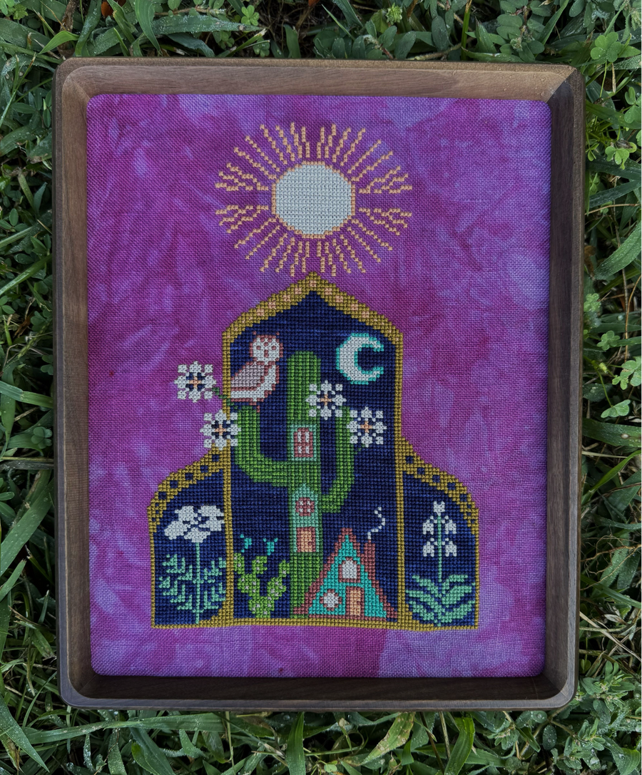 A framed needlepoint canvas featuring a whimsical house with a cactus, owl, moon, and sun on a purple tie-dye background, placed outdoors on green grass.
