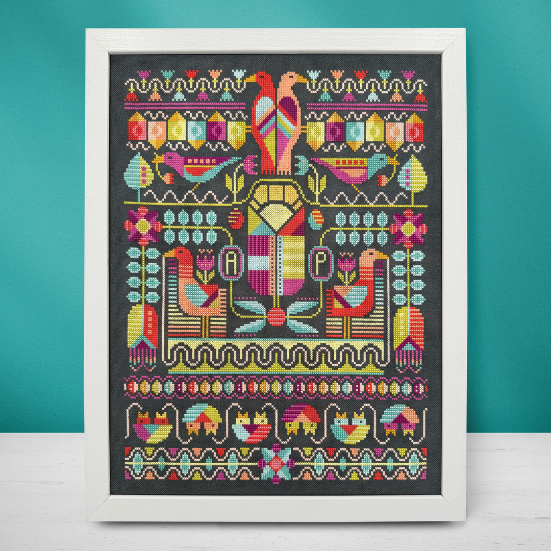 A framed cross-stitch artwork featuring a colorful symmetrical folkloric design with birds, floral motifs, and decorative borders on a dark fabric background, set against a teal wall and white surface.