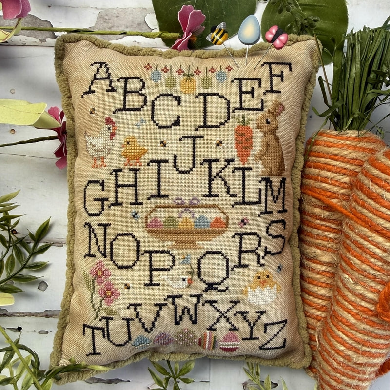 Decorative pillow with cross-stitched alphabet and farm-themed illustrations on a wooden surface with plants.