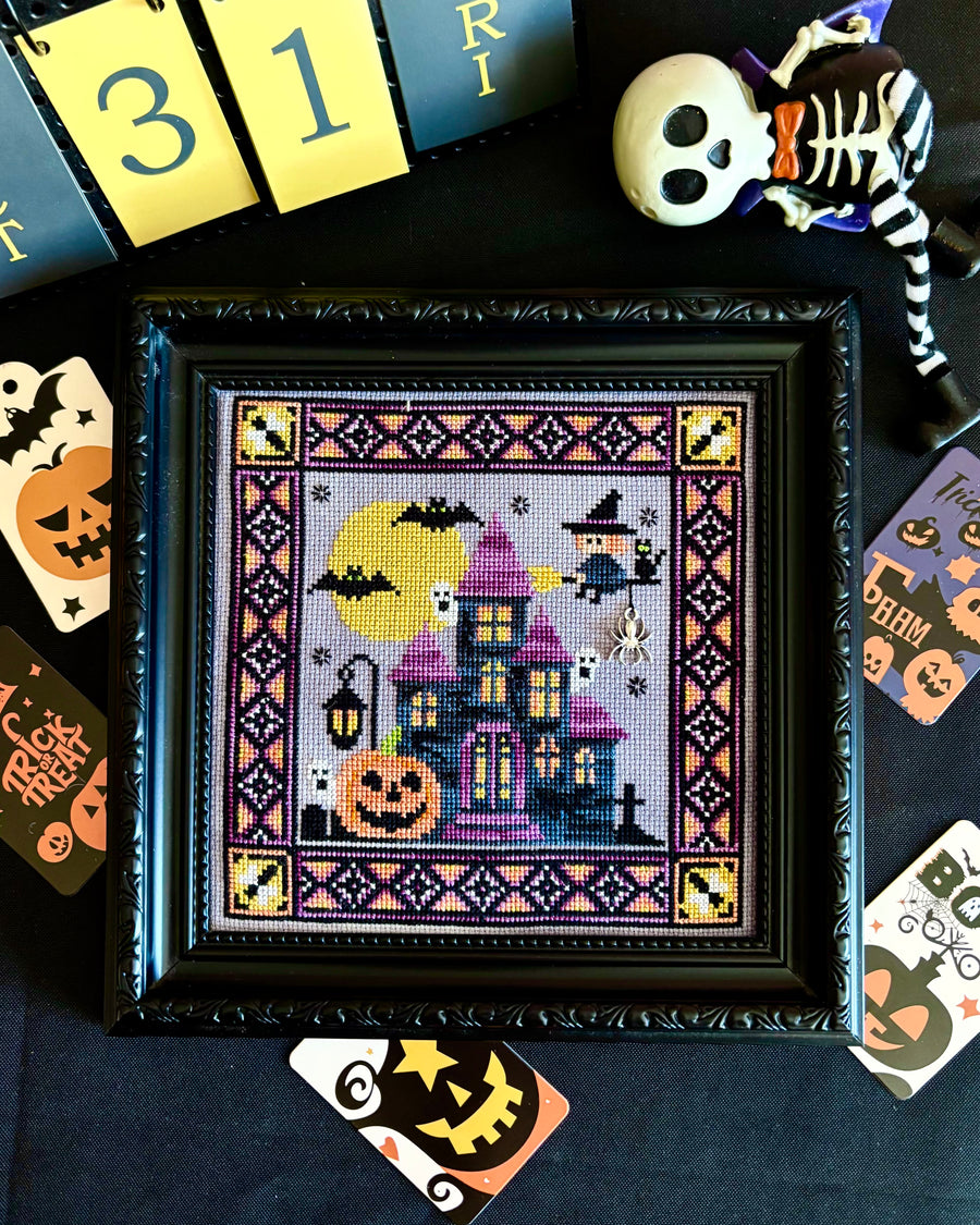 A framed Halloween cross-stitch artwork featuring a haunted purple house, jack-o'-lantern, bats, and a witch under a full moon, displayed on a dark surface with seasonal props including cards, a skeleton decoration, and a calendar.