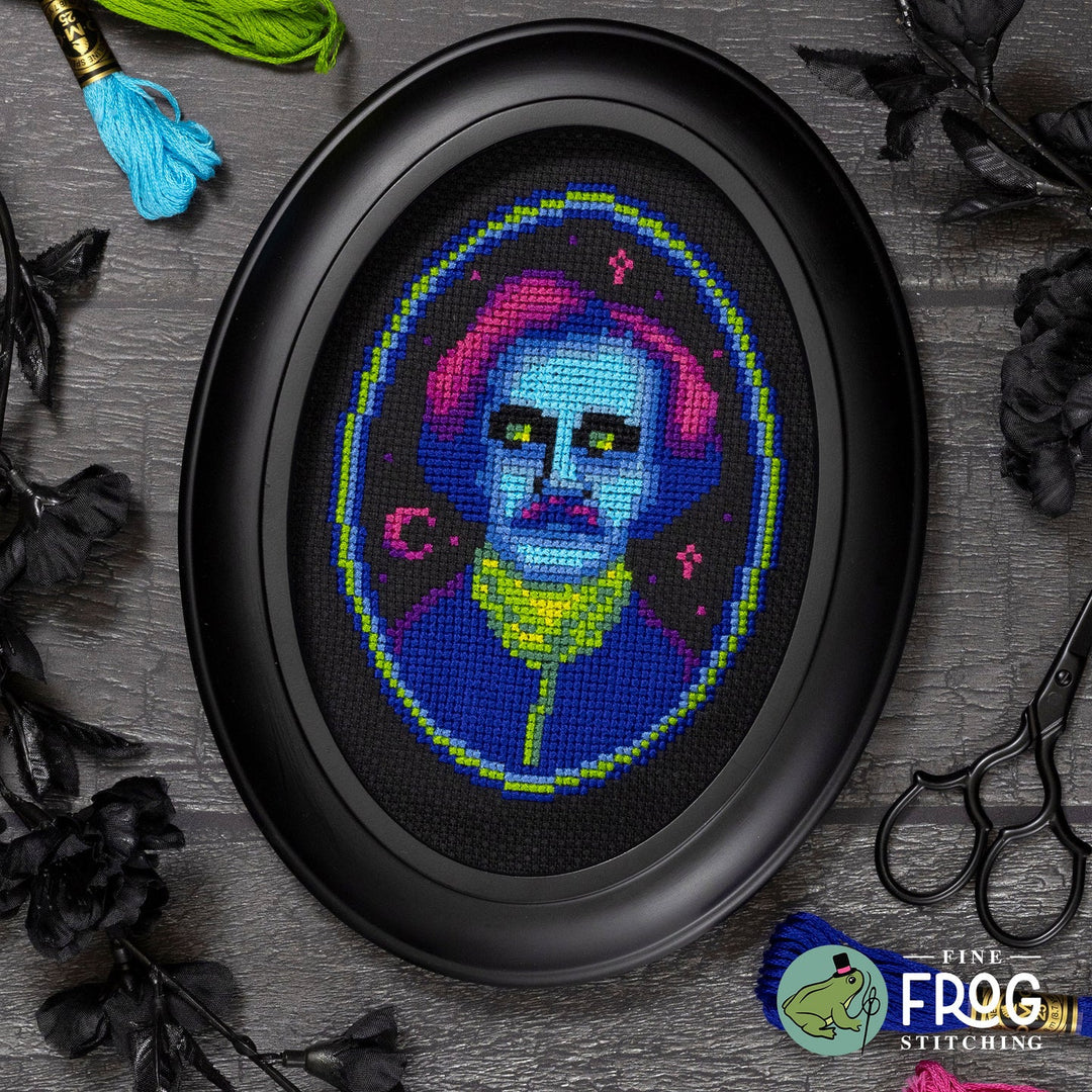 An oval framed cross-stitch design featuring a stylized blue-faced figure with pink hair and a yellow collar, displayed on a dark gray textured surface with decorative black flowers, scissors, and tinsel. A 'Fine Frog Stitching' logo is visible in the bottom right corner.