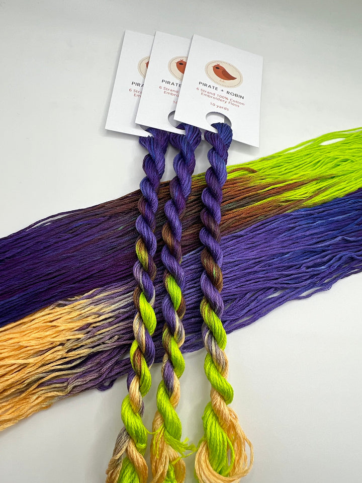 Three braided skeins of embroidery floss with attached white cards, displayed over a fan of matching loose threads. The floss and thread are multicolor, showing gradients of purple, yellow, brown, and white.