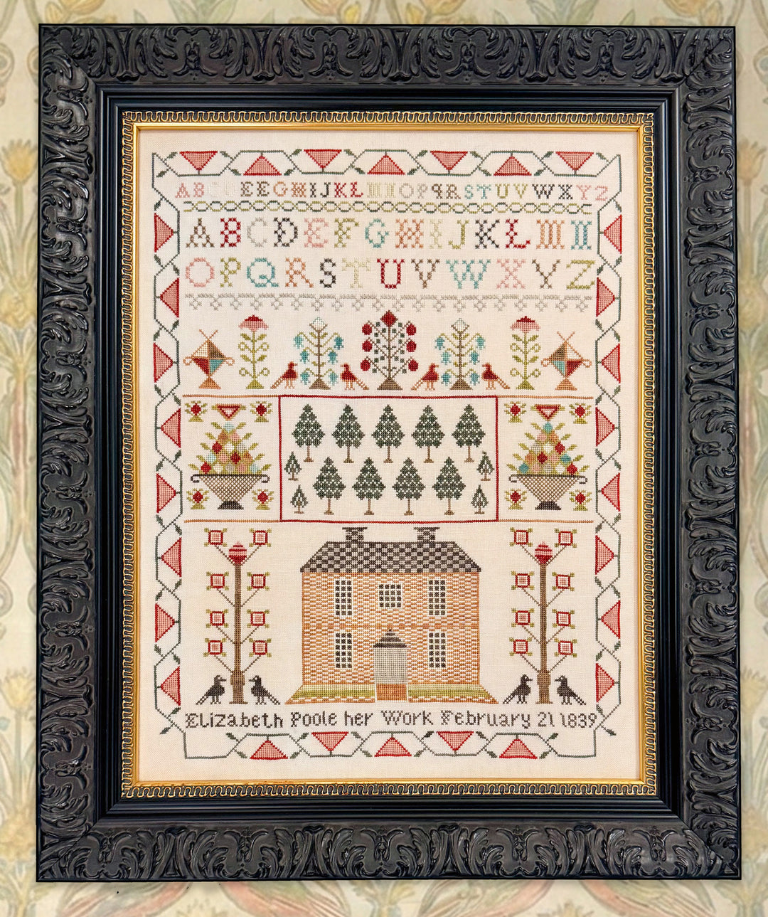 Decorative cross-stitch pattern with alphabet, trees, and a house on a beige background, framed in black.