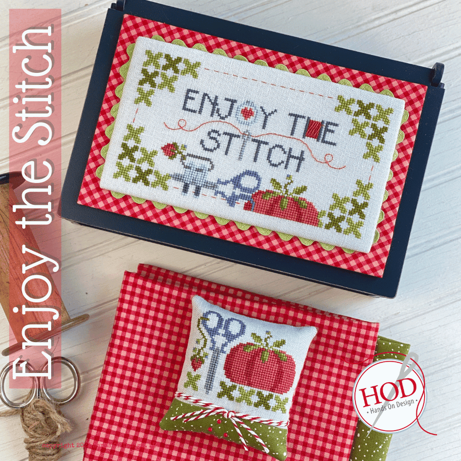 Cross-stitched fabric with 'Enjoy the Stitch' text on a red checkered background, surrounded by sewing tools.