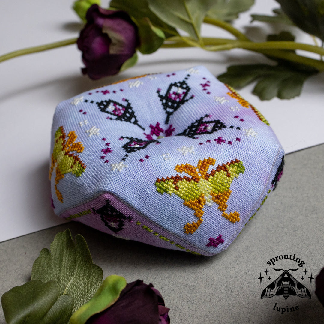 A small hexagonal fabric pillow with embroidered butterfly and floral patterns in yellow, orange, purple, and black on a pale lavender background, styled with dried flowers on a white and gray surface. The Sprouting Lupine brand logo is visible in the bottom right corner.