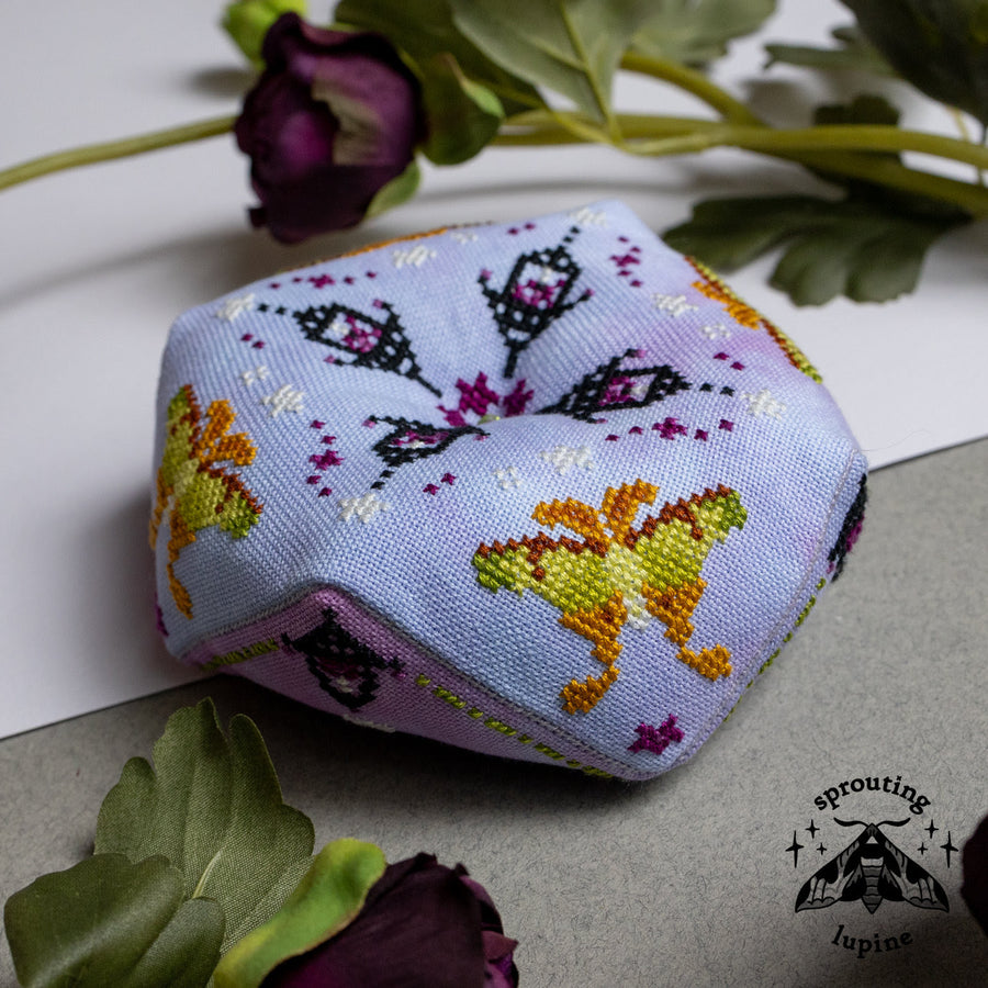 A small hexagonal fabric pillow with embroidered butterfly and floral patterns in yellow, orange, purple, and black on a pale lavender background, styled with dried flowers on a white and gray surface. The Sprouting Lupine brand logo is visible in the bottom right corner.