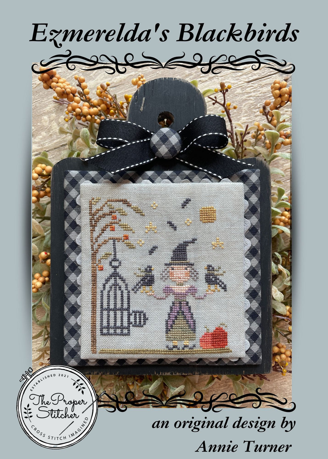 Cross-stitched design of a witch with a blackbird and birdcage, framed by decorative elements.