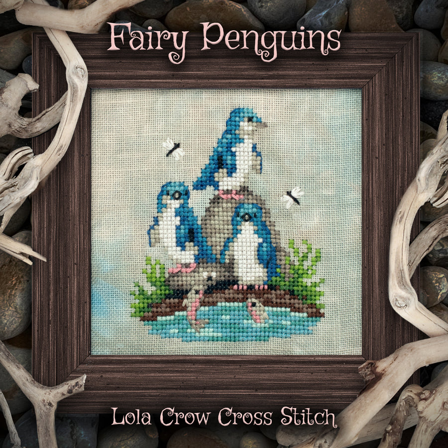 A framed cross stitch artwork titled "Fairy Penguins" by Lola Crow Cross Stitch, showing three blue-and-white penguins on rocks by a body of water. The framed piece is centered against a background of driftwood and stones.