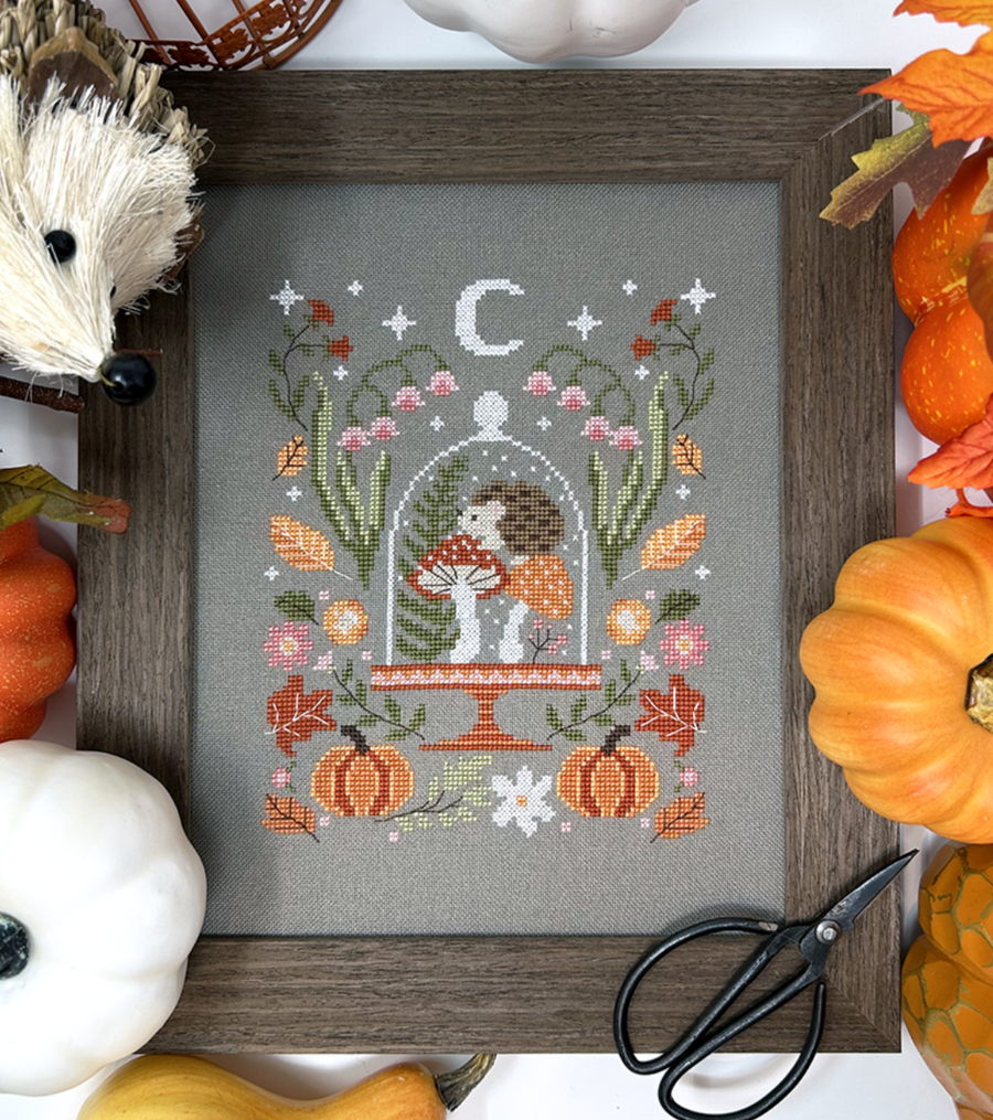 Cross-stitched piece with autumnal design in a wooden frame, surrounded by pumpkins and scissors.