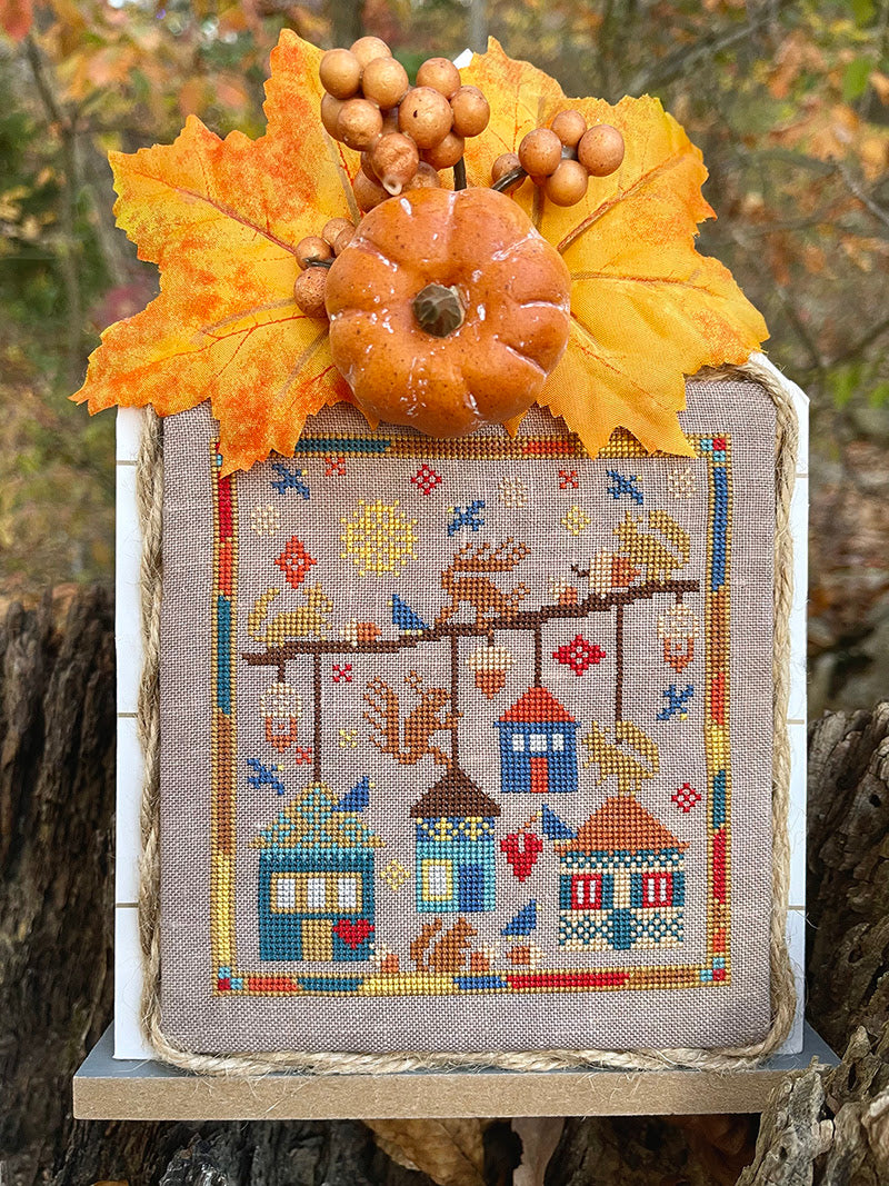 Cross-stitched scene with autumn elements including a pumpkin and leaves on a textured background.