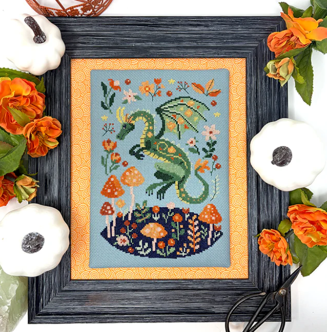 Framed cross-stitched artwork of a green dragon among flowers and mushrooms, surrounded by decorative pumpkins and flowers.
