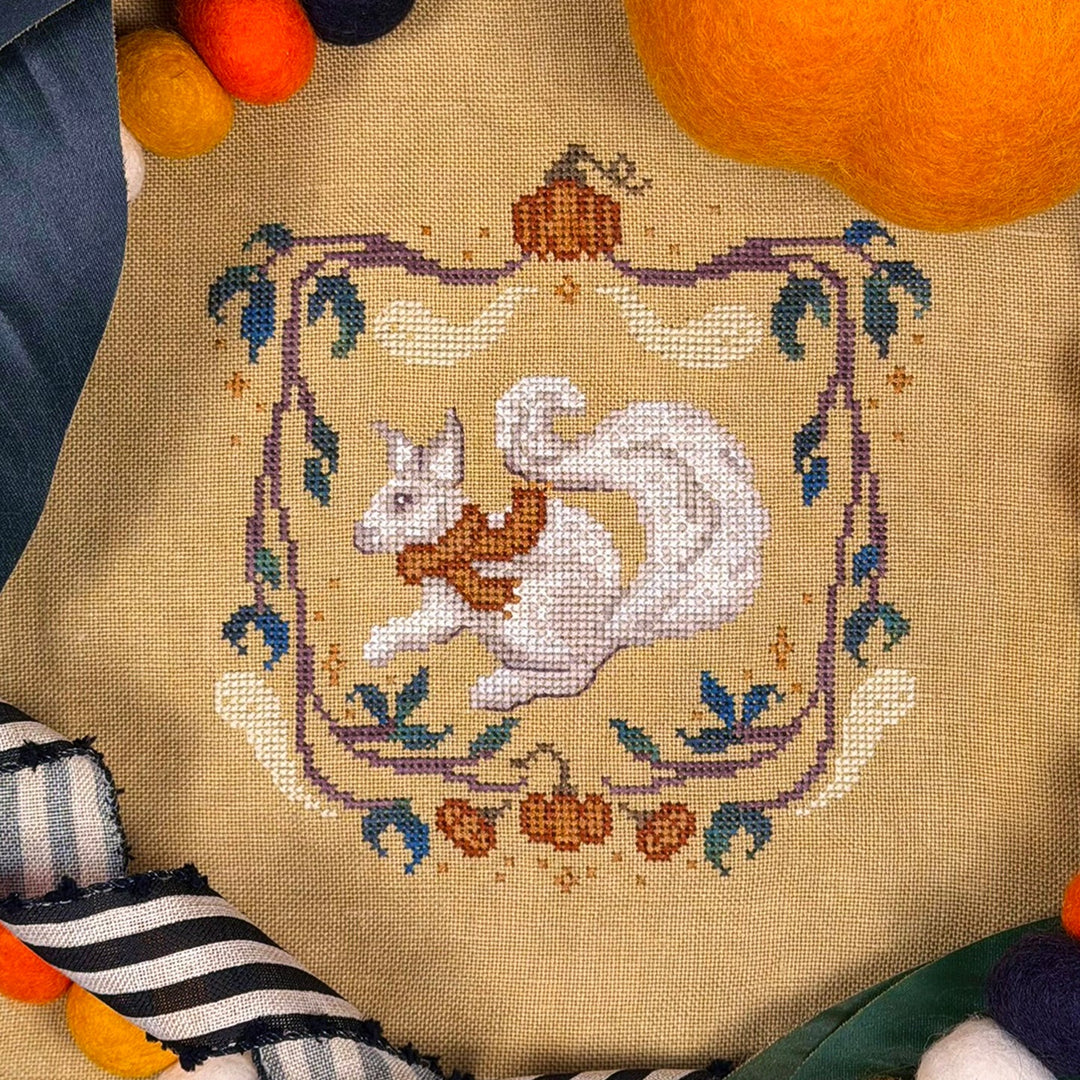 A close-up of a finished cross-stitch design featuring a white squirrel wrapped in an orange scarf inside an autumn-themed border with pumpkins and leaves, stitched on a beige fabric background with decorative yarn and fabric props around it.