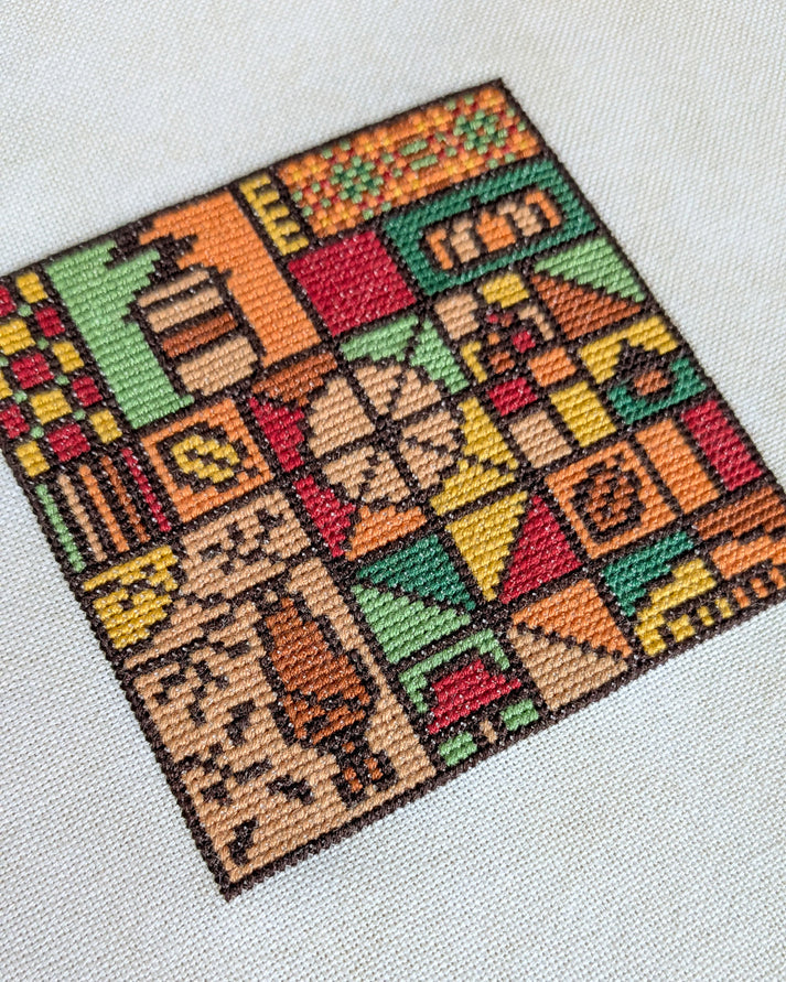 A square cross-stitch sampler featuring a colorful patchwork design of houses, trees, and geometric shapes, displayed on a light beige textured fabric background.