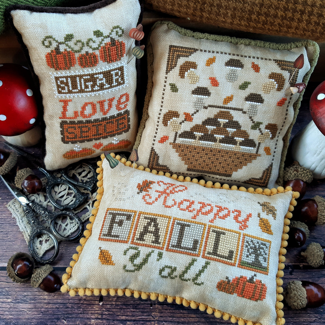 Three cross-stitched pillows with autumn-themed designs on a wooden surface.