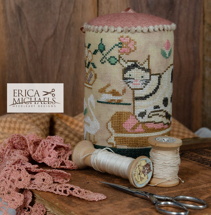 Decorative pin cushion with cross-stitched design of a cat, surrounded by yarn, scissors, and lace on a wooden surface.