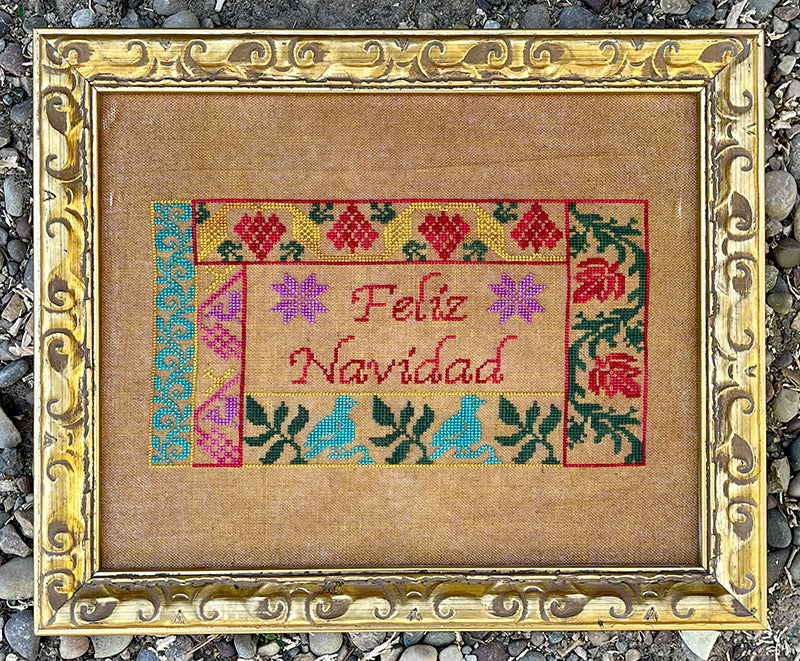 Cross-stitched 'Feliz Navidad' design in a decorative gold frame on a textured surface.