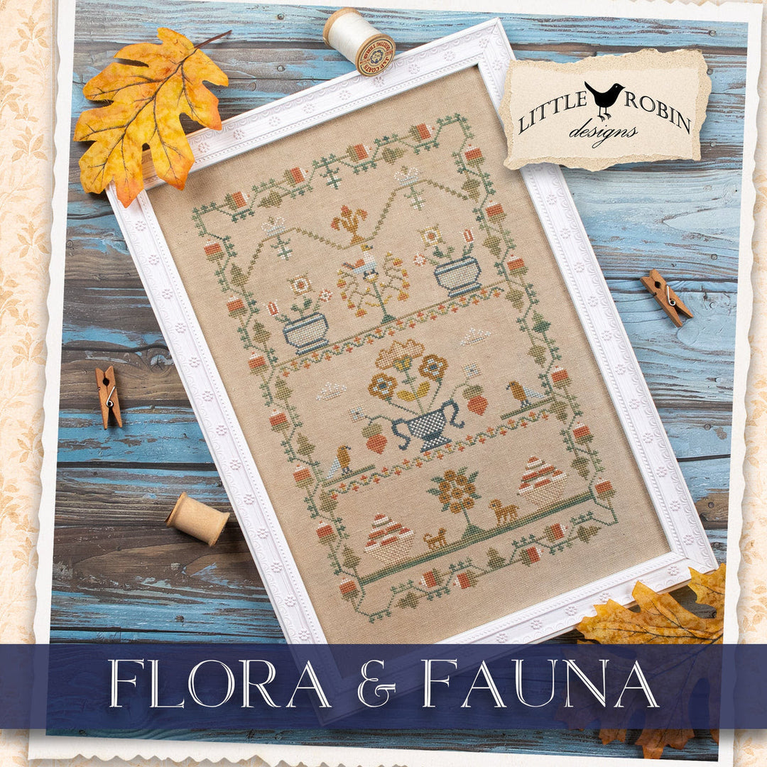 A framed cross-stitch design titled 'Flora & Fauna' by Little Robin Designs, featuring a beige fabric background with embroidered botanical and animal motifs in muted earthy tones, styled on a rustic blue wooden surface with autumn leaves, spools of thread, and wooden clothespins.