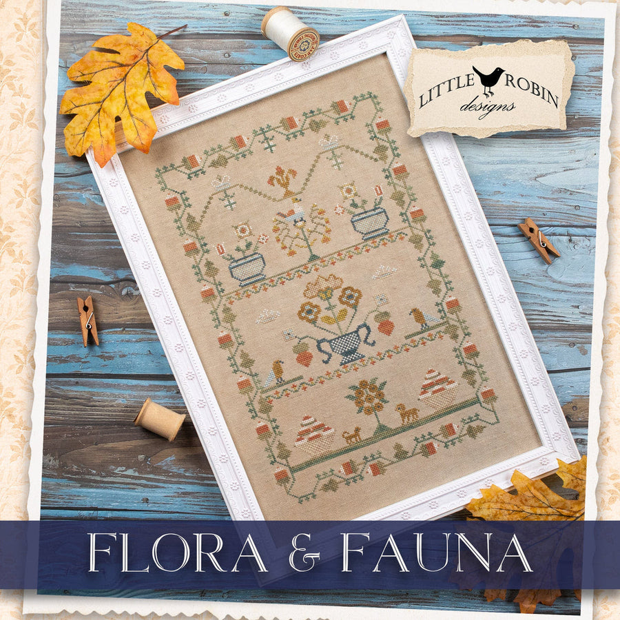 A framed cross-stitch design titled 'Flora & Fauna' by Little Robin Designs, featuring a beige fabric background with embroidered botanical and animal motifs in muted earthy tones, styled on a rustic blue wooden surface with autumn leaves, spools of thread, and wooden clothespins.