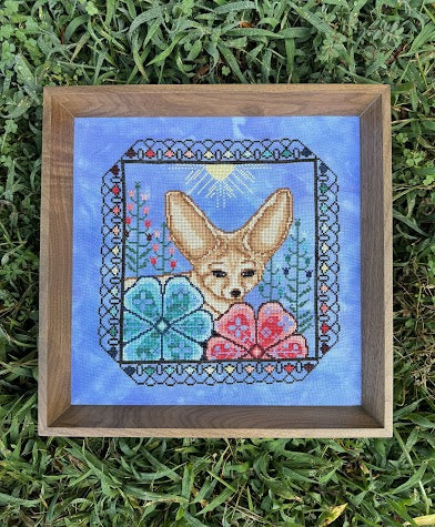 A framed cross-stitch artwork featuring a fennec fox surrounded by flowers on a blue background, placed on green grass.