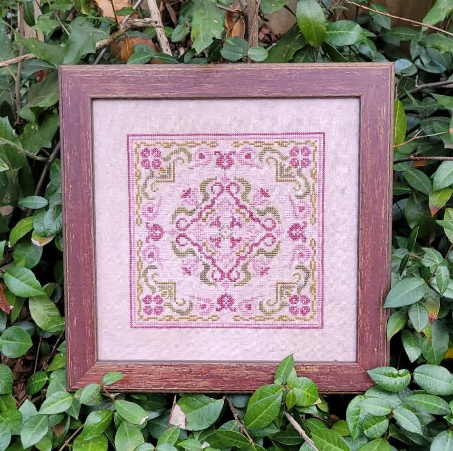 Framed cross-stitched pattern on a pink background, surrounded by green leaves.