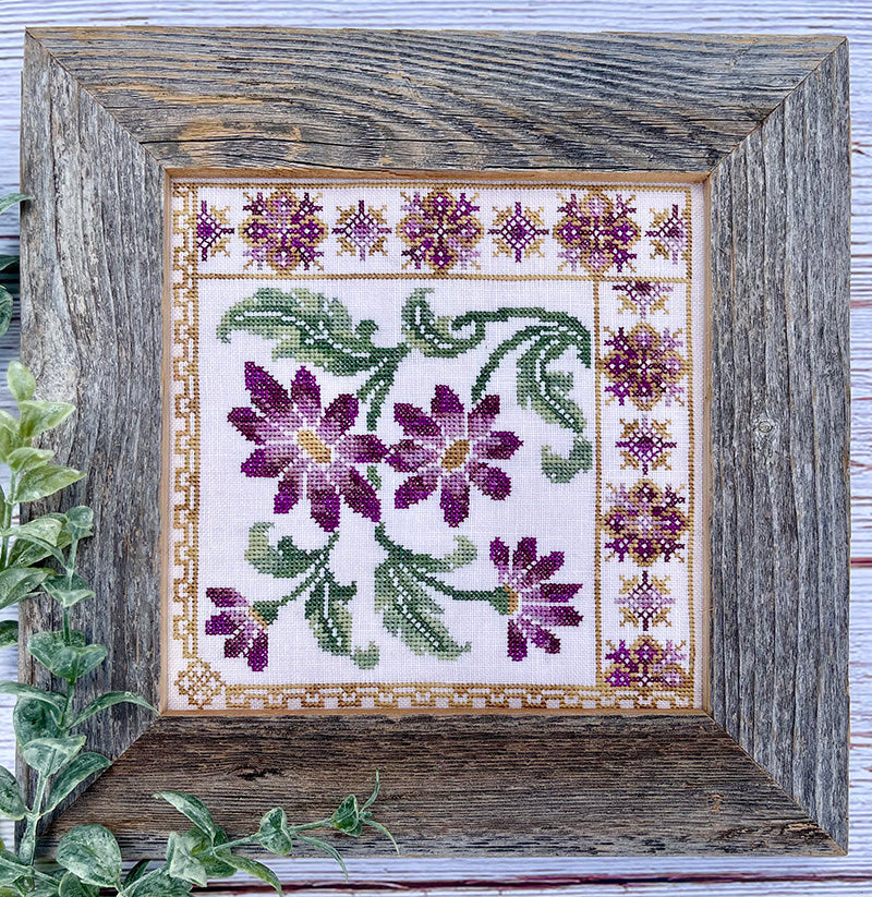 Framed cross-stitched floral design with purple flowers and green leaves on a white background.