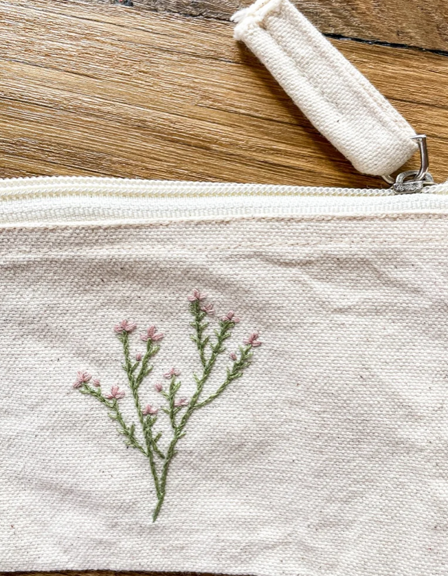 A close-up of a cream-colored canvas zipper pouch featuring an embroidered green and pink wildflower design on the front, resting on a wooden surface.