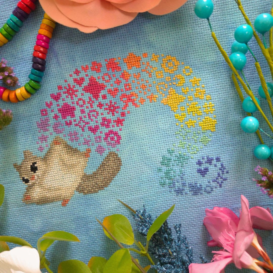 A close-up of a cross-stitch or needlework textile featuring a small beige animal with a rainbow made of colorful flower-like stitches. The scene is styled with flowers, greenery, and bead strands around the stitched design.