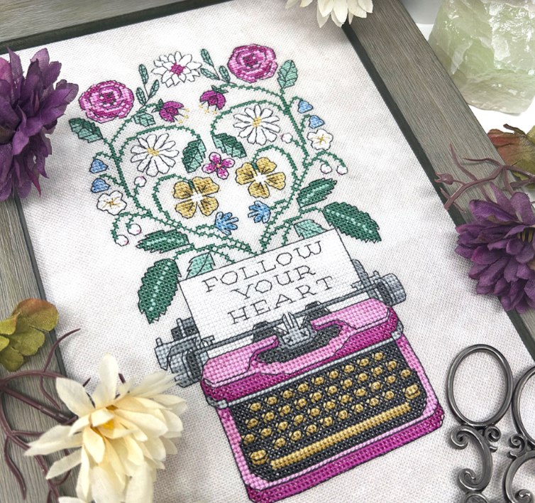 Cross-stitched design of a typewriter with flowers and 'Follow Your Heart' text, surrounded by flowers and decorative items.