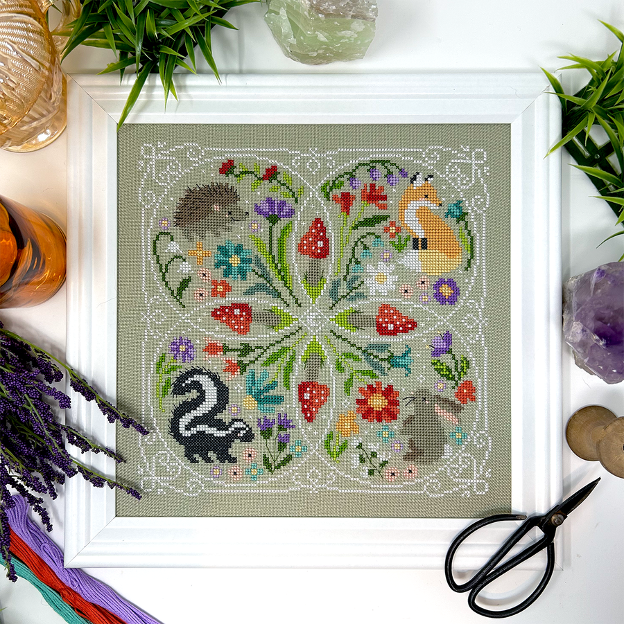 A framed cross-stitch needlework piece depicting a forest scene with mushrooms, flowers, and animals such as a fox, hedgehog, skunk, and rabbit, surrounded by a white border with green plants and crafting accessories.