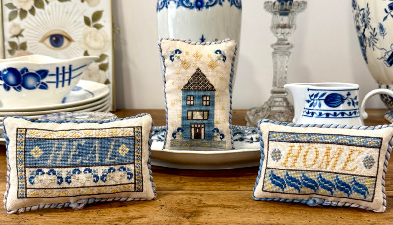 Three decorative embroidered throw pillows arranged on a wooden table with blue-and-white ceramic dishes in the background. The left pillow says "HEAL," the center pillow shows a small house, and the right pillow says "HOME.