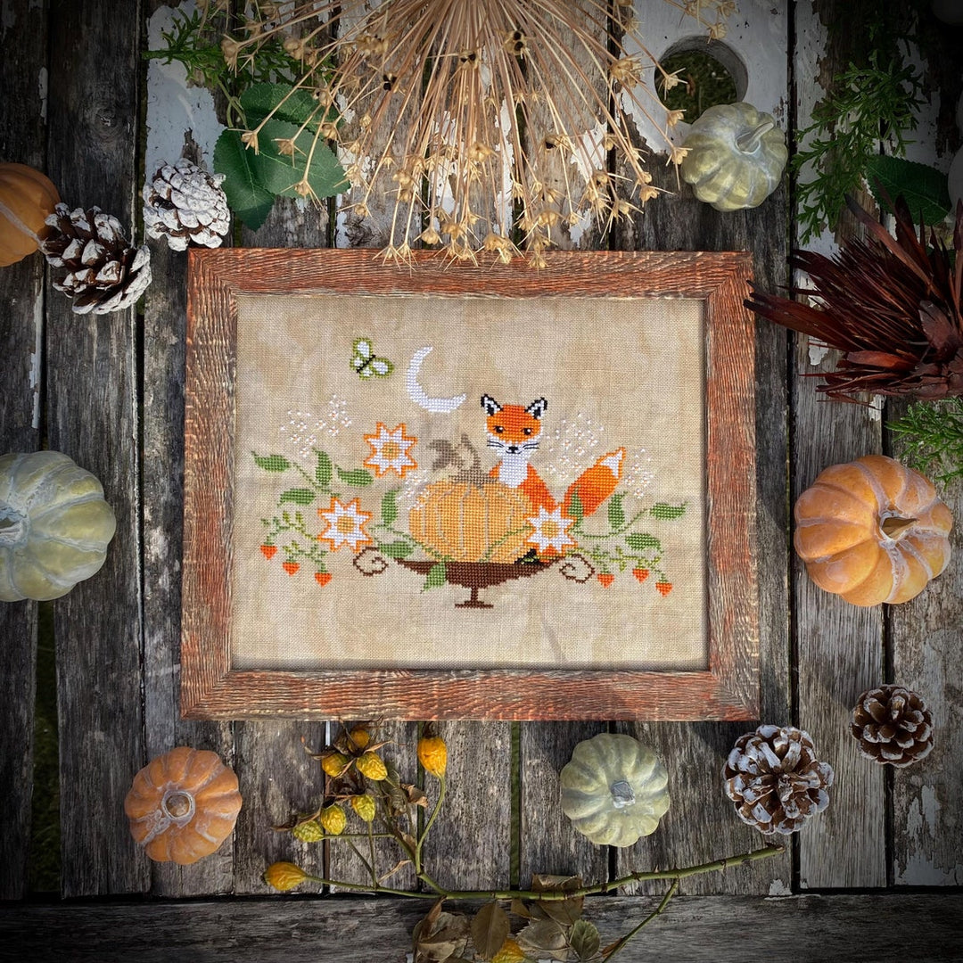 Framed cross-stitched picture of a fox and pumpkins on a wooden surface with autumn decorations.