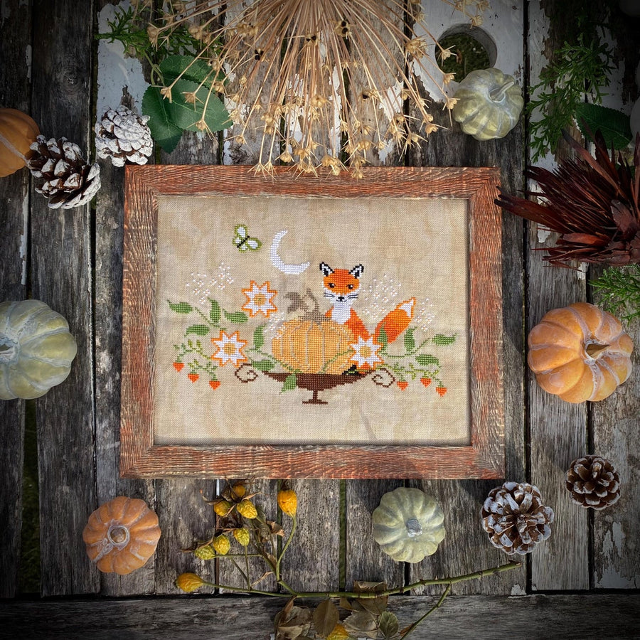 Framed cross-stitched picture of a fox and pumpkins on a wooden surface with autumn decorations.