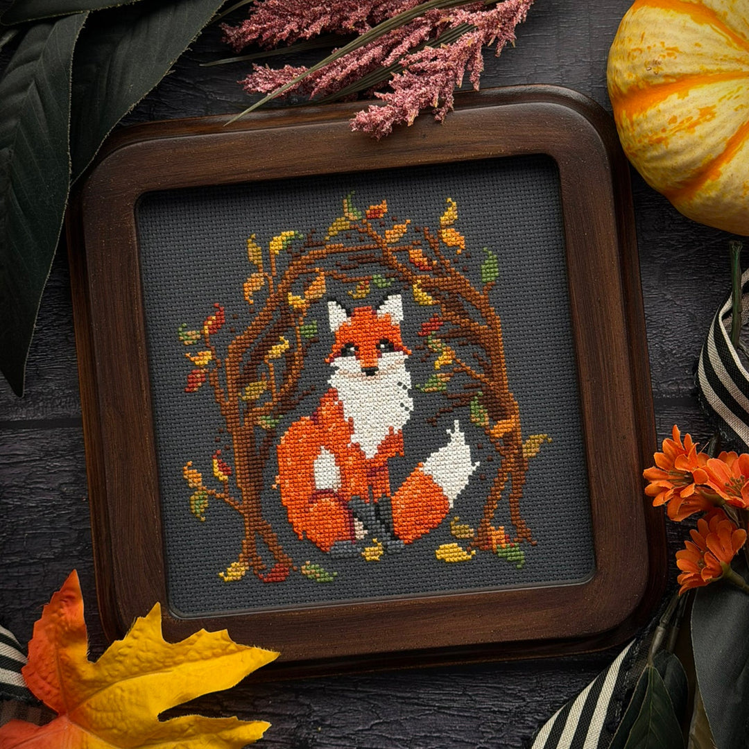 A framed cross-stitch artwork featuring an orange and white fox surrounded by autumn leaves and branches, set in a dark wood frame and styled with fall leaves, dried flowers, and a small pumpkin on a dark textured surface.