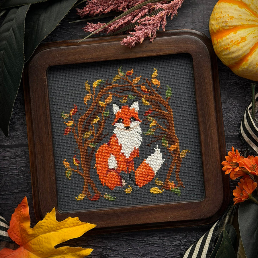 A framed cross-stitch artwork featuring an orange and white fox surrounded by autumn leaves and branches, set in a dark wood frame and styled with fall leaves, dried flowers, and a small pumpkin on a dark textured surface.