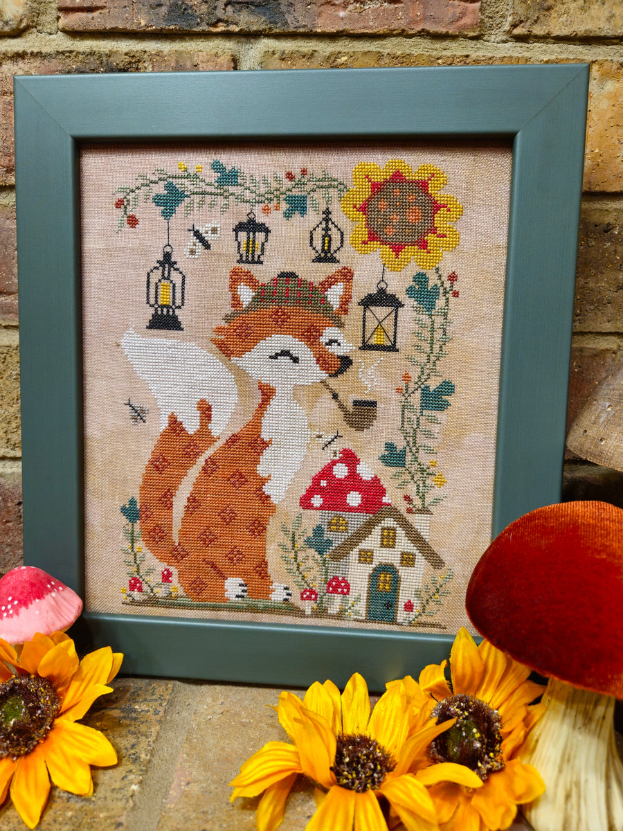 A framed cross-stitch artwork featuring a fox, mushrooms, lanterns, and sunflowers, displayed against a brick wall with decorative flowers in the foreground.