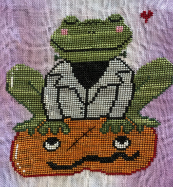 A section of a needlepoint canvas with a green frog wearing a white shirt and sitting on an orange background, surrounded by purple linen.