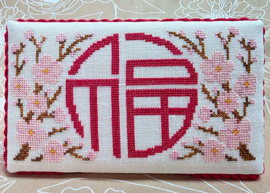 A rectangular cross-stitch cushion featuring a large red Chinese character for 'fortune' surrounded by pink cherry blossoms on a white fabric background.