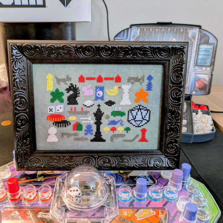 Framed cross-stitch pattern of colorful game pieces on a table with gaming supplies.