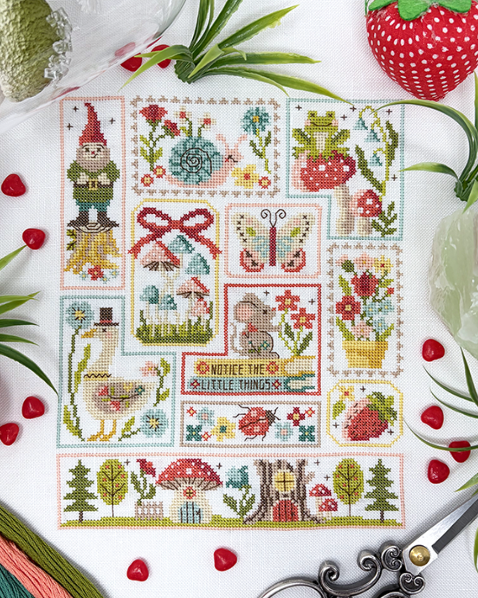 A cross-stitch pattern sheet featuring various whimsical garden and nature motifs, including a gnome, strawberry, flowers, a butterfly, a bird, and a row of trees and mushrooms. The sheet is styled in a flat-lay composition with embroidery floss, scissors, and decorative strawberries on a white background.