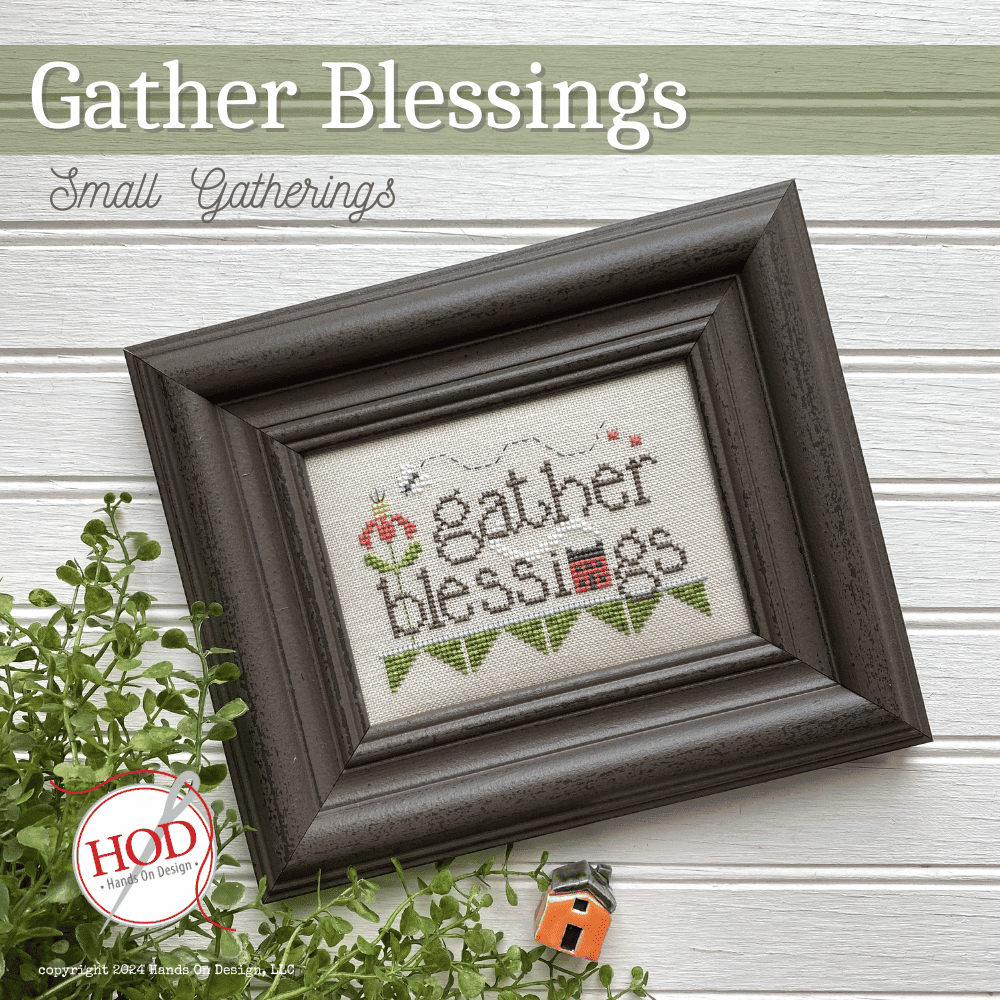 Framed cross-stitched artwork with 'gather blessings' text on a wooden surface.