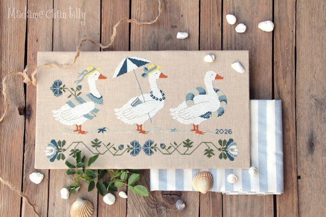 A mounted cross stitch canvas featuring three white decorative geese with blue, gray, and orange details on a tan background, laid flat on a rustic wooden surface with shells, twine, and folded striped fabric around it. 