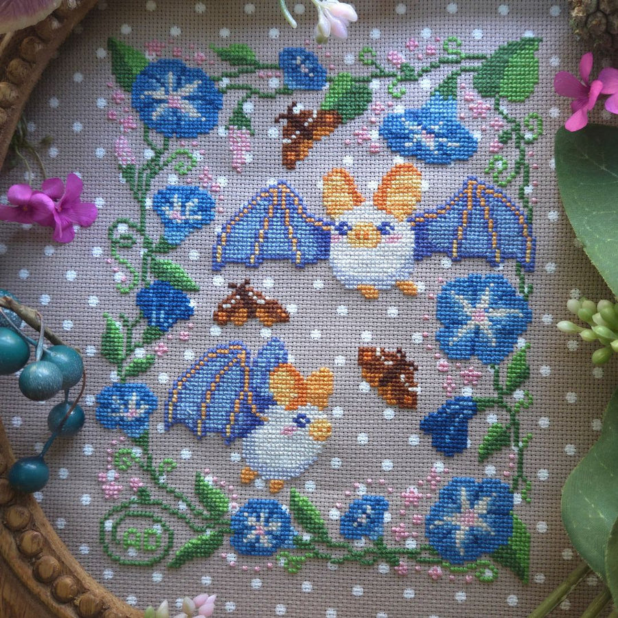 A close-up of a circular embroidered cross-stitch piece featuring two small bats with blue wings, surrounded by blue flowers, green vines, pink blossoms, and brown leaves on a white polka-dot fabric background. The hoop is framed by wood and small decorative flowers.