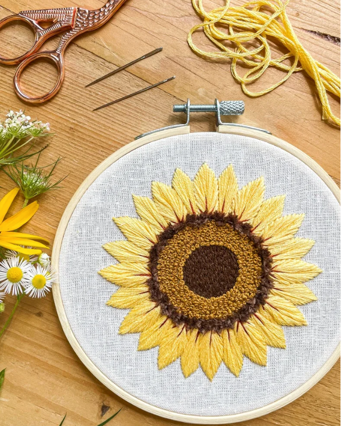 A completed sunflower embroidery piece in a round wooden hoop, displayed on a wooden table with embroidery tools, yellow thread, flowers, and copper-colored scissors.