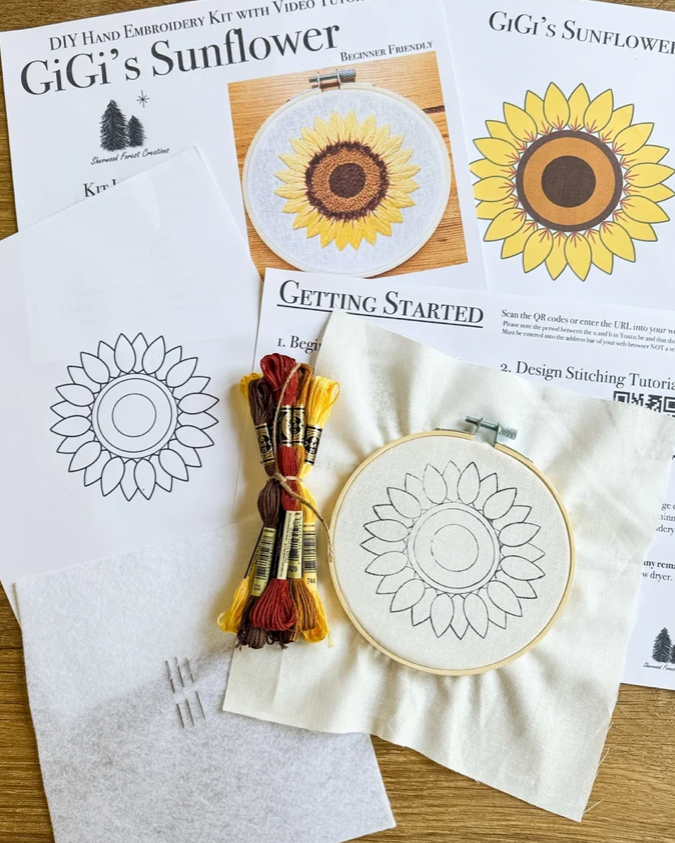 Sunflower DIY hand embroidery kit laid out on a wooden surface, including printed pattern sheets, embroidery floss in yellow, red, and brown, metal needles, and two fabric hoops with sunflower embroidery designs.
