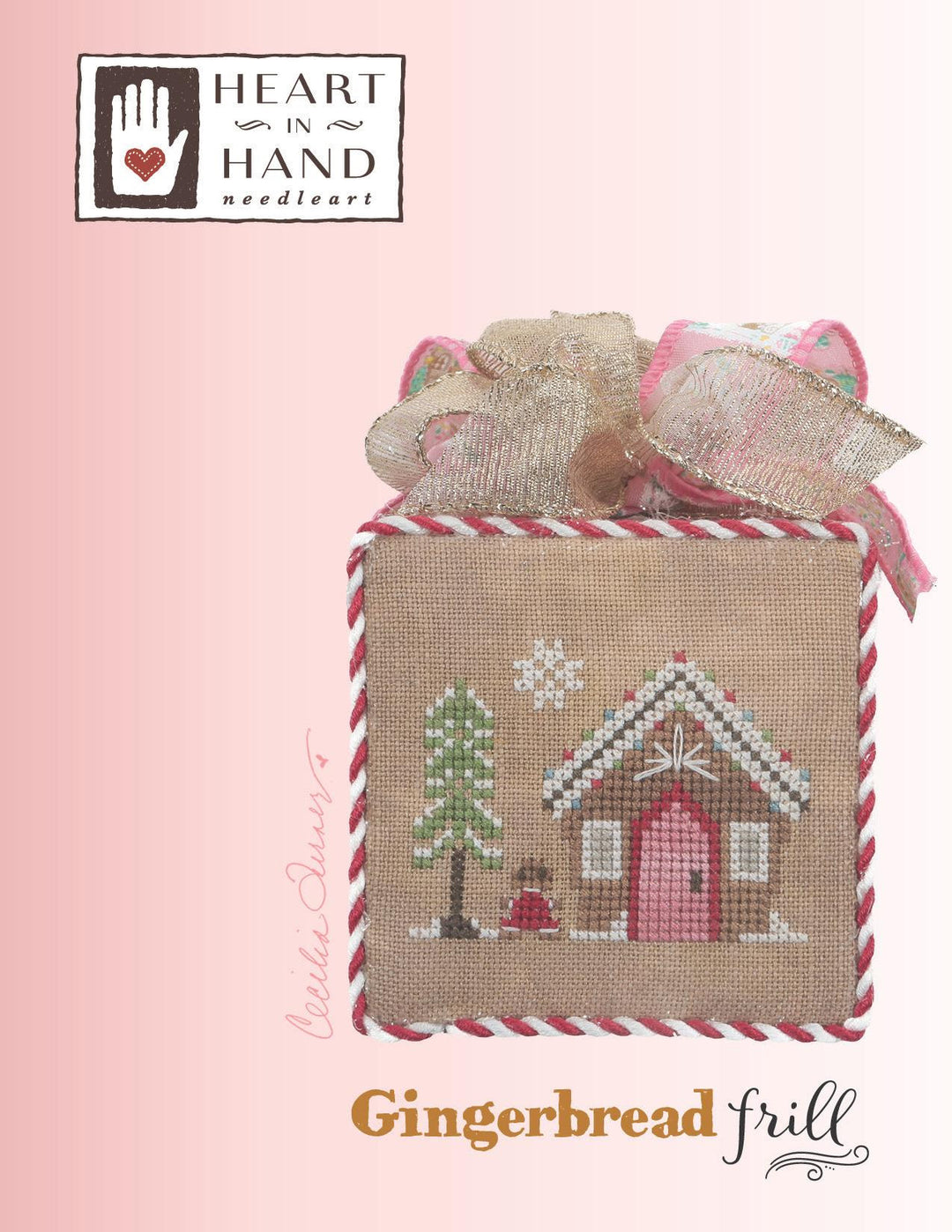 Heart in Hand Needleart 'Gingerbread Frill' kit showing a finished cross-stitch design of a gingerbread house and tree on a burlap fabric square, tied with a ribbon bow, on a pink gradient background with the brand logo and product name.