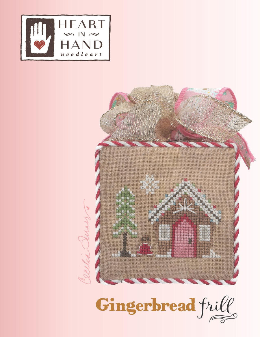 Heart in Hand Needleart 'Gingerbread Frill' kit showing a finished cross-stitch design of a gingerbread house and tree on a burlap fabric square, tied with a ribbon bow, on a pink gradient background with the brand logo and product name.