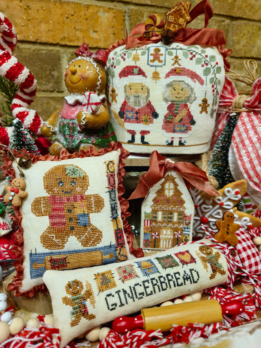 A collection of hand-embroidered Christmas gingerbread-themed home decor items, including pillows, a hanging ornament, and a cylindrical pillow reading 'GINGERBREAD', arranged with candy canes, a gingerbread doll, and holiday decorations against a brick wall background.
