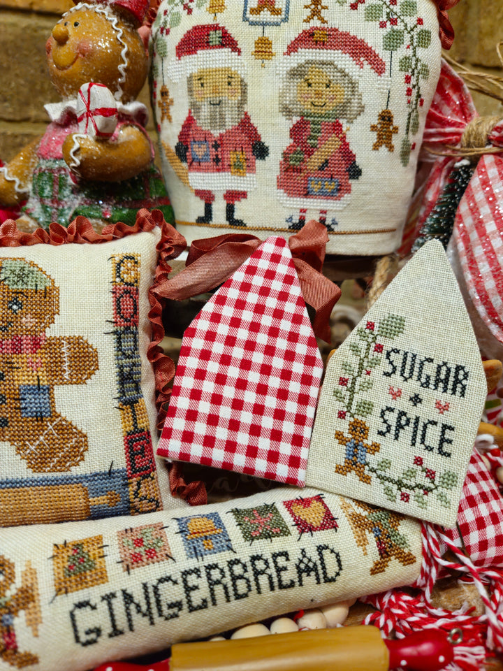 A collection of Christmas-themed cross-stitch needlework pieces, including a gingerbread cookie pillow, a red and white gingham house, a 'Sugar Spice' house, a 'Gingerbread' scroll, and a Santa and gingerbread woman sampler, arranged together with a gingerbread man figurine.