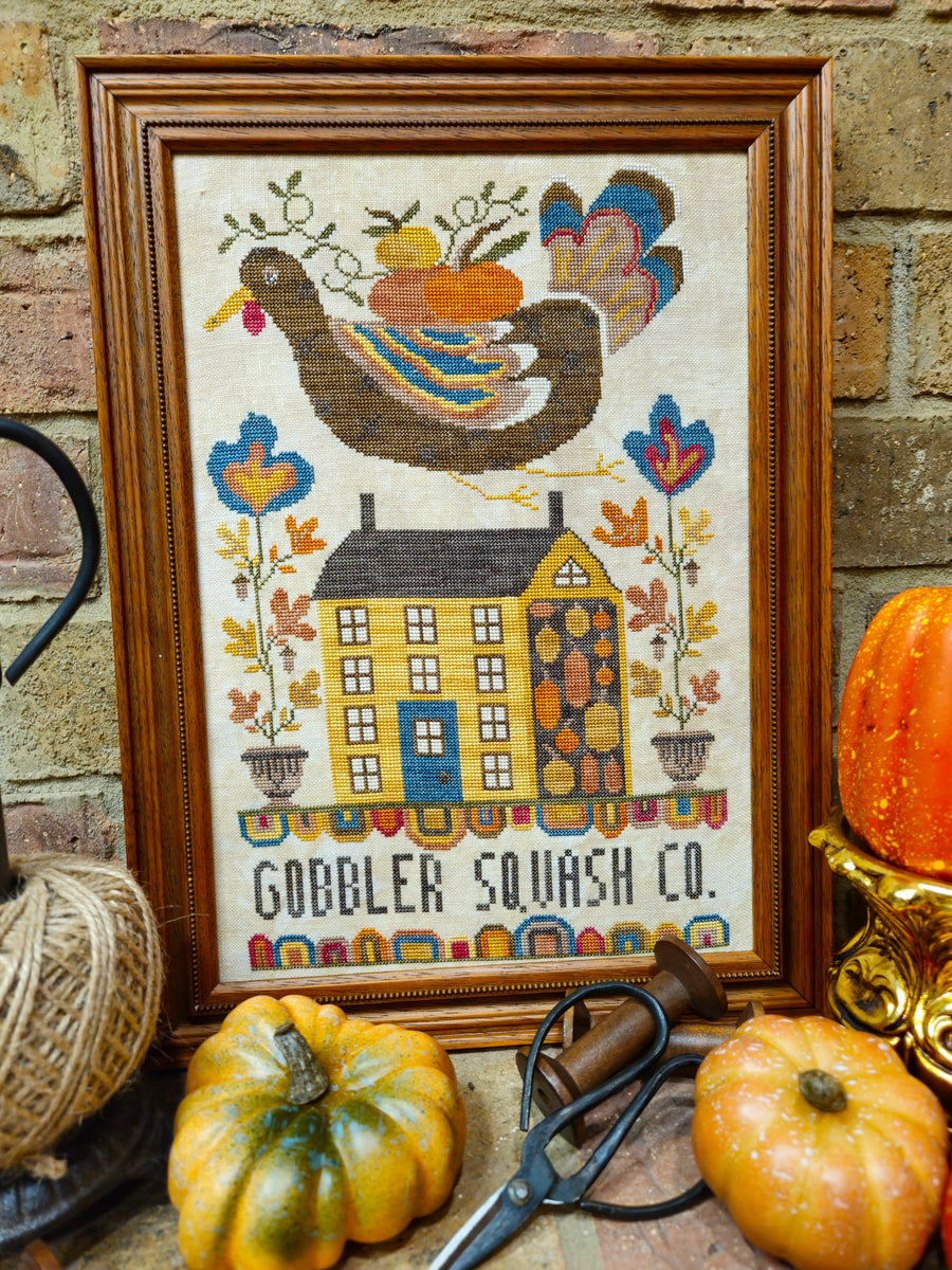 A framed cross-stitch artwork featuring a house with the text 'GOBLER SQUASH CO.' and a large turkey carrying pumpkins, styled with small pumpkins, scissors, twine, and a brick wall background.