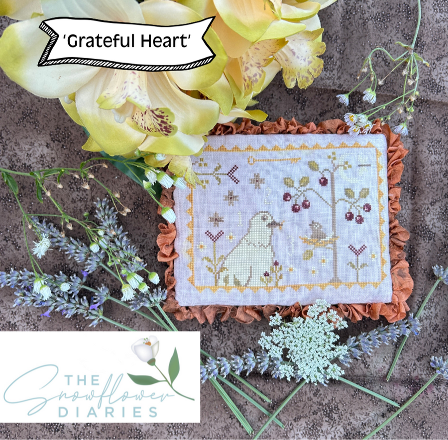 Decorative pillow with cross-stitch design surrounded by flowers and plants, featuring 'Grateful Heart' and 'The Snowflower Diaries' branding.